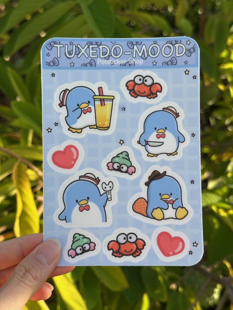May include: A sheet of stickers featuring a blue penguin wearing a sailor hat and bow tie in various poses. The stickers also include a red crab, a green sea slug, and a heart. The sheet is on a blue background with white stars and the text "TUXEDO-MOOD" and "Potsticker Shop".