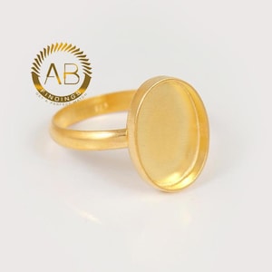 Puede incluir: Un anillo base de color dorado con un engaste ovalado liso diseñado para cabujones. La superficie metálica tiene un acabado mate. Un logotipo circular con el texto AB FINDINGS GET A PERFECT MATCH es visible en la esquina superior izquierda.