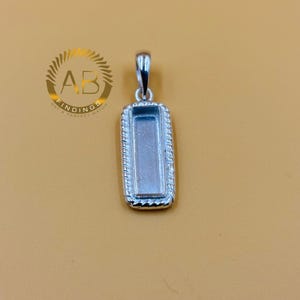 May include: A silver-colored rectangular pendant setting with a rope-like border. The pendant has a loop at the top for a chain and a recessed area to hold a stone or other element. The background is a solid tan color.