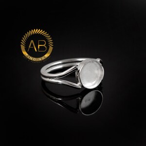 K&ouml;nnte beinhalten: Ein silberfarbener Metallring mit einer kreisf&ouml;rmigen Fassung f&uuml;r einen Cabochon oder eine Einlage. Die Ringschiene teilt sich in zwei d&uuml;nne Arme, die mit der runden, flachen Schale verbunden sind. Ein goldenes Logo mit dem Text AB FINDINGS ist sichtbar.