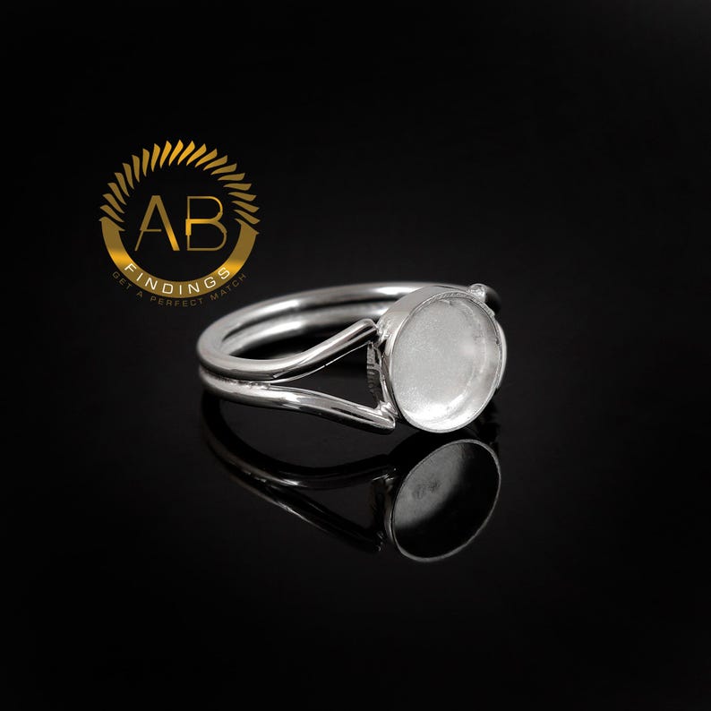 K&ouml;nnte beinhalten: Ein silberfarbener Metallring mit einer kreisf&ouml;rmigen Fassung f&uuml;r einen Cabochon oder eine Einlage. Die Ringschiene teilt sich in zwei d&uuml;nne Arme, die mit der runden, flachen Schale verbunden sind. Ein goldenes Logo mit dem Text AB FINDINGS ist sichtbar.