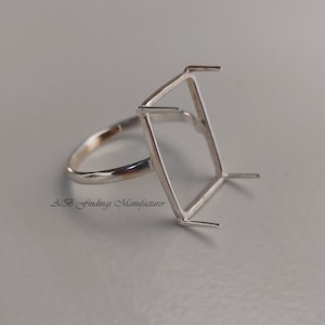 Puede incluir: Un engaste de anillo plateado con una parte superior rectangular, diseñado para sostener una piedra preciosa. El anillo tiene una banda simple y cuatro garras en cada esquina del engaste. El texto "AB Findings Manufacturer" es visible.