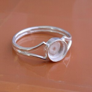 K&ouml;nnte beinhalten: Ein silberfarbener Metallring mit einem geteilten Ringschienen-Design, das in einer kreisf&ouml;rmigen Fassung zusammenl&auml;uft. Die Fassung ist leer und f&uuml;r einen Stein oder Cabochon vorgesehen, wobei der Ring eine glatte, polierte Oberfl&auml;che aufweist.
