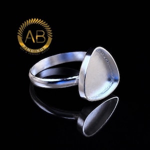 May include: A silver-toned metal ring blank featuring a triangular bezel setting designed for stone or cabochon placement. The ring has a smooth, polished band and a flat, recessed triangular cup. The logo AB FINDINGS GET A PERFECT MATCH is visible.