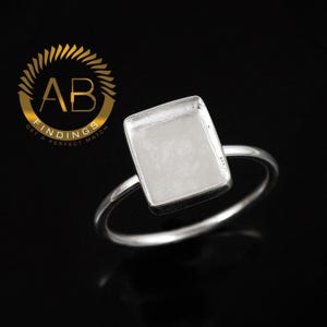 May include: A silver ring with a square bezel setting. The ring has a simple band and a rectangular setting, perfect for a custom design. The ring is displayed against a black background. The logo "AB FINDINGS GET A PERFECT MATCH" is in the upper left corner.