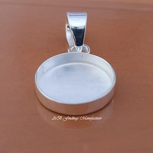 Puede incluir: Un colgante plateado con un bisel redondo y un lazo para una cadena. El engaste está diseñado para sostener una piedra redonda o cabujón. El texto "AB Findings Manufacturer" está en la parte inferior.