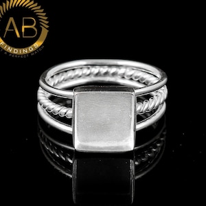 May include: A silver ring featuring a central square bezel setting for a stone or inlay. The band consists of three distinct strands, with two outer smooth wires and a central twisted rope-style wire, creating a layered and textured appearance.