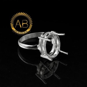 Puede incluir: Un engaste de anillo plateado con un centro de forma ovalada y cuatro garras. El anillo tiene una banda simple y un engaste detallado diseñado para sujetar una piedra preciosa. El logotipo "AB FINDINGS" está en la esquina superior izquierda.