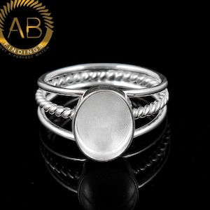 Puede incluir: Un anillo de plata con un engaste ovalado central diseñado para una piedra. La banda consta de tres hilos distintos, con el hilo central que muestra una textura de cuerda trenzada, mientras que los dos hilos exteriores son lisos y sencillos.