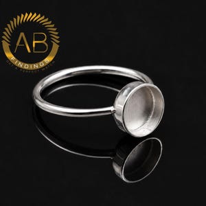 May include: A silver-toned metal ring blank featuring a circular bezel cup setting designed for jewellery making. The ring has a smooth, polished band and a plain, empty cup ready for a stone or cabochon. The logo AB FINDINGS is visible.