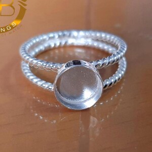 Könnte beinhalten: Ein silberfarbener Ring mit einem doppelten Band in gedrehter Seiloptik. In der Mitte des Rings befindet sich eine kreisförmige Fassung mit flachem Boden, die für einen Stein oder Cabochon vorgesehen ist und eine glatte, polierte Oberfläche aufweist.