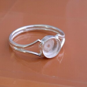 K&ouml;nnte beinhalten: Ein silberfarbener Ring mit einem geteilten Ringschienen-Design, das in einer kreisf&ouml;rmigen Fassung zusammenl&auml;uft. Die Fassung ist leer und daf&uuml;r vorgesehen, einen Stein oder Cabochon aufzunehmen. Das Metall hat eine glatte Oberfl&auml;che.