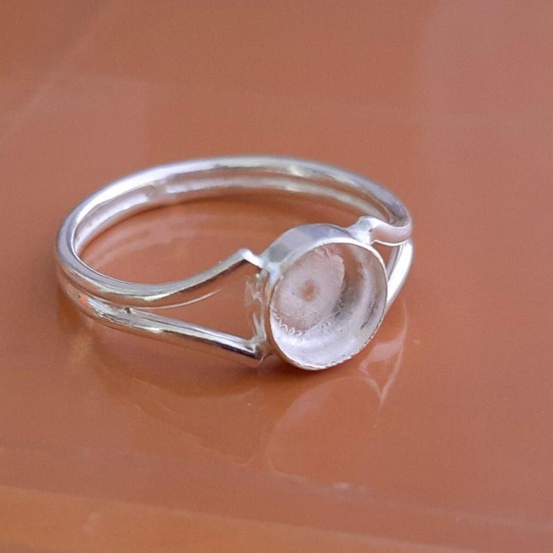 K&ouml;nnte beinhalten: Ein silberfarbener Ring mit einem geteilten Ringschienen-Design, das in einer kreisf&ouml;rmigen Fassung zusammenl&auml;uft. Die Fassung ist leer und daf&uuml;r vorgesehen, einen Stein oder Cabochon aufzunehmen. Das Metall hat eine glatte Oberfl&auml;che.