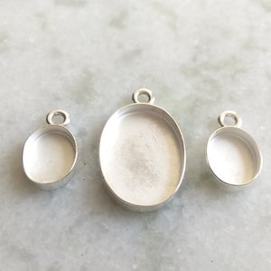 May include: Three silver oval pendant trays with a loop at the top for attaching a chain. The trays are different sizes.