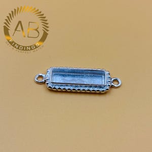 May include: A silver-coloured rectangular jewellery finding with a beaded border and two loops. The centre is a recessed area for setting a stone or other embellishment. The logo "AB FINDINGS GET A PERFECT MATCH" is in the upper left corner.