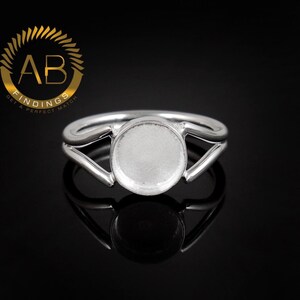 K&ouml;nnte beinhalten: Ein silberfarbener Ring mit einer kreisf&ouml;rmigen Fassung f&uuml;r einen Cabochon oder Stein. Die Ringschiene teilt sich in zwei Arme, die in die zentrale runde Fassung &uuml;bergehen. Ein kreisf&ouml;rmiges Logo mit dem Text AB FINDINGS ist zu sehen.