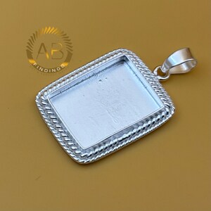 May include: A silver rectangular pendant setting with a rope-style border. The pendant has a loop for a chain and a recessed area for a stone or design. The logo "AB FINDINGS" is visible in the upper left corner.