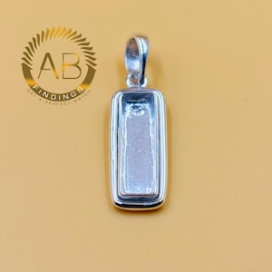 May include: A silver rectangular pendant with a loop for a chain. The pendant has a recessed area in the centre. The background is a solid, light brown colour. The pendant is likely for jewellery making.