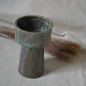 Può includere: Un vaso in ceramica con una sezione superiore cilindrica larga caratterizzata da linee verticali incise e una base affusolata. L'oggetto presenta una smaltatura multicolore nei toni del blu profondo, marrone e verde acqua, con finitura liscia.