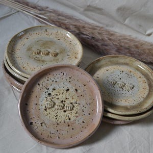 Może przedstawiać: Stos okrągłych ceramicznych talerzy z nakrapianym szkliwem w odcieniach beżu, kremu i delikatnego różu. Każdy talerz posiada mały, wypukły wzór przedstawiający trzy babeczki umieszczone na środku powierzchni, o gładkiej i błyszczącej fakturze.