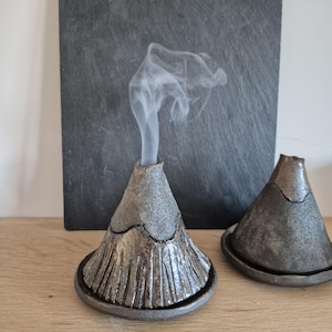 May include: Two cone-shaped ceramic incense burners with a textured, dark metallic finish. The left burner features vertical ridges at the base and emits a thin trail of white smoke from its top opening. Both pieces sit on matching circular trays.