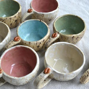 Op de afbeelding: Een verzameling van zeven handgemaakte keramische mokken met onregelmatige, organische vormen. Elke mok heeft een gespikkelde crèmekleurige buitenkant en een gladde, effen gekleurde binnenkant in pastelroze, lichtblauw of saliegroen, met een koffieboon.
