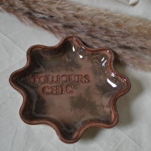 Könnte beinhalten: Eine sternförmige Keramikschale mit einer glänzenden, gesprenkelten braunen Glasur. In der Mitte der Schale befindet sich der geprägte Schriftzug "TOUJOURS CHIC" in Großbuchstaben. Die Ränder der Schale sind gewellt und erhöht, was eine sternförmige Silhouette ergibt.