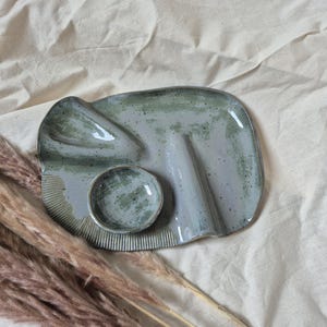 May include: A handmade ceramic serving tray with a glossy sage green and grey speckled glaze. The tray features an irregular organic shape, a small attached circular bowl, two raised ridges, and a textured ribbed pattern along one curved edge.