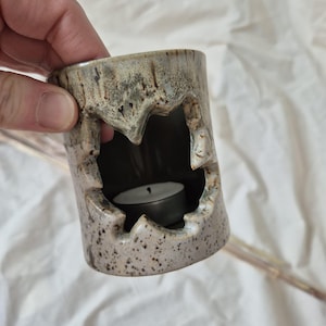 Może przedstawiać: Cylindryczny ceramiczny świecznik z nakrapianą beżowo-szarą glazurą. Zewnętrzna strona posiada duży, poszarpany i nieregularny otwór, odsłaniający ciemne wnętrze, w którym znajduje się mała, okrągła, biała świeczka typu tea light.