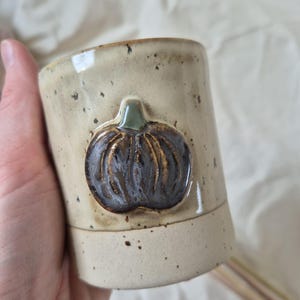 Może przedstawiać: Cylindryczny kubek ceramiczny z nakrapianą, kremową glazurą. Na zewnętrznej stronie znajduje się wypukły, trójwymiarowy relief przedstawiający ciemnofioletową dynię z małą zieloną łodyżką, umieszczony w górnej części kubka.