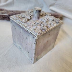 Puede incluir: Una caja cuadrada de cerámica con tapa que presenta un asa cilíndrica central. La superficie tiene un acabado de esmalte moteado en rosa pálido y blanco con puntos oscuros. La tapa está decorada con una densa disposición de flores de cerámica.
