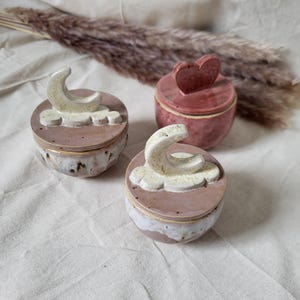 Op de afbeelding: Drie kleine keramische potten met deksel en een glanzende afwerking. Twee potten hebben een crème-kleurig handvat in de vorm van een maansikkel en wolk op het deksel, terwijl de derde pot een roze hartvormig handvat heeft.
