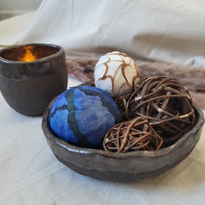 Peut inclure: Un bol en céramique brun foncé contenant une sphère bleue avec des lignes noires, une sphère blanche avec des motifs bruns et deux sphères en brindilles tressées. À côté du bol, une tasse en céramique brun foncé contient une lumière chaude.