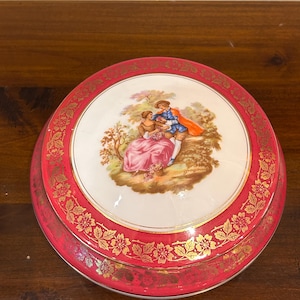 Op de afbeelding: Ronde, rood en goudkleurige sieradendoos met deksel. Het deksel toont een geschilderde scène van een stel in een tuin. De doos heeft een gouden bloemenpatroon langs de rand. De doos is waarschijnlijk van keramiek.