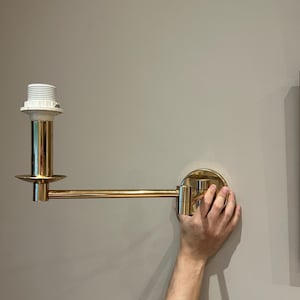 Può includere: Una lampada da parete color ottone con braccio regolabile. La lampada presenta un portalampada bianco e una base circolare. Il braccio si estende orizzontalmente. La lampada è montata su una parete color neutro.