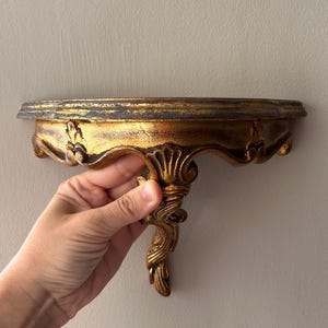 May include: A semi-circular wall shelf featuring a distressed gold finish with dark undertones. The shelf has a flat top surface and a decorative, carved support bracket underneath with ornate, scrolling leaf-like details and a pointed base.