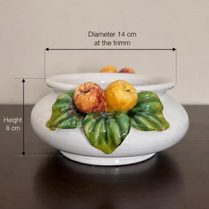 Peut inclure: Un bol en céramique blanche orné d'un relief décoratif représentant deux pommes et des feuilles vertes sur le devant. Le bol mesure 8 cm de hauteur et 14 cm de diamètre au niveau du bord. La surface est lisse et brillante, avec des fruits et des feuilles peints aux couleurs naturelles.