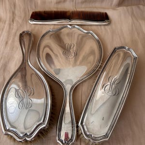 Op de afbeelding: Een kapset bestaande uit een zilverkleurige handspiegel, twee haarborstels en een kam. Elk stuk heeft een gepolijste metalen afwerking met een decoratief gegraveerd monogram in het midden. De kam heeft een rand in een donkerbruine schildpadstijl.