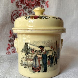 May include: A cream-colored ceramic canister with a matching lid, featuring a decorative floral border and a detailed illustration of people in traditional clothing at an outdoor market scene. The cylindrical container has two small side handles.