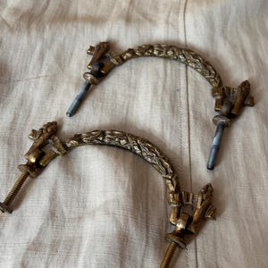 May include: Two matching antique brass drawer pulls featuring a curved, arch-shaped handle with a detailed leaf and vine relief pattern. Each pull includes two threaded metal bolts with nuts for mounting, showing a weathered, aged patina finish.
