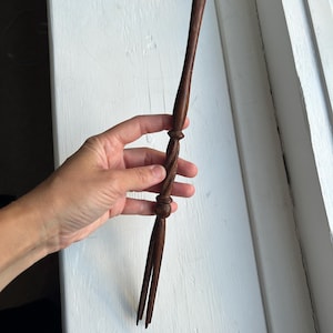 Puede incluir: Un tenedor de madera marrón oscuro con un mango retorcido y dos púas. El tenedor se sostiene en una mano sobre un fondo blanco. El mango tiene un diseño decorativo con una forma bulbosa en el medio.