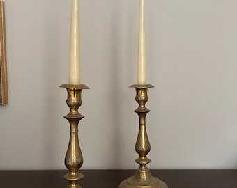 Puede incluir: Dos candelabros altos de latón pulido con intrincados patrones florales grabados en la base y el tallo. Cada candelabro sostiene una vela cónica de color blanco roto. El metal tiene un acabado dorado cálido y reflectante con base circular.