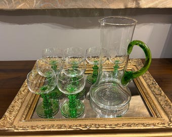 May include: A vintage glassware set featuring a clear glass pitcher with a green handle and a set of matching wine glasses with green stems. The set is displayed on a gold-framed mirrored tray, creating an elegant presentation.