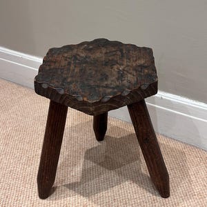 Puede incluir: Un taburete de madera de color marrón oscuro con un asiento hexagonal que presenta bordes festoneados y una superficie texturizada. El taburete se apoya sobre tres patas cilíndricas gruesas que se estrechan ligeramente hacia abajo, mostrando una veta de madera visible.