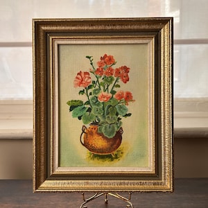 May include: A framed oil painting on canvas depicting a cluster of orange-red geranium flowers with green leaves in a textured copper-colored pot. The artwork is enclosed in an ornate, metallic gold-toned frame with a light-colored inner border.