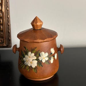 May include: A wooden jar with a lid, decorated with hand-painted white flowers and green leaves. The jar has two small handles and a faceted knob on the lid. The jar is brown and sits on a black surface.