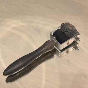 Op de afbeelding: Een antieke prentmaker met een donkerhouten handvat en een metalen frame. De wals heeft een zwarte roller en een kleinere, getextureerde roller. De tool is ontworpen om inkt gelijkmatig op een oppervlak aan te brengen.