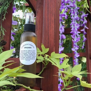 May include: A clear glass bottle with a brown cap and label that reads "Ayurvedic Hair Oil Organic". The bottle is filled with a golden liquid and is sitting in front of a wooden fence with purple flowers.