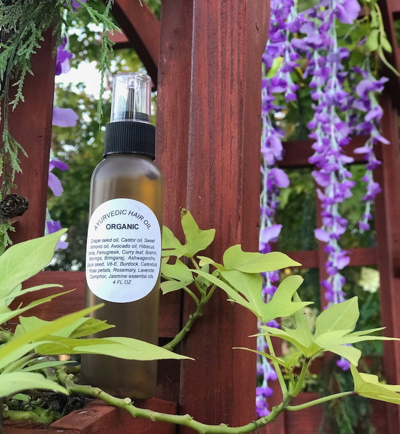May include: A clear glass bottle with a brown cap and label that reads "Ayurvedic Hair Oil Organic". The bottle is filled with a golden liquid and is sitting in front of a wooden fence with purple flowers.