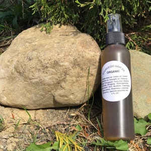 May include: A brown glass bottle with a spray nozzle, labeled "AYURVEDIC HAIR OIL ORGANIC" and listing ingredients including grape seed oil, castor oil, sweet almond oil, avocado oil, hibiscus, fenugreek, curry leaf, brahmi, amla, bringaraj, ashwagandha, black seed, vit-e, burdock, calendula, rose petals, rosemary, lavender, camphor, jasmine essential oils. The bottle is sitting on a rock and green grass.